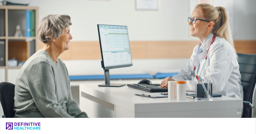 An older woman talking to a younger woman in a white lab coat sitting behind a desk with a computer in front of her