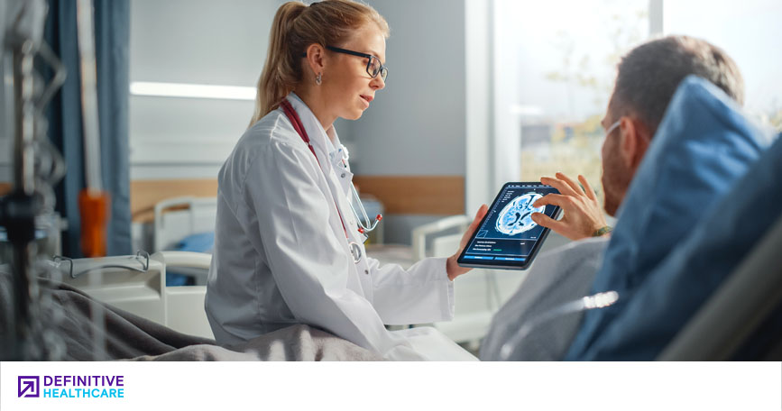 A female doctor sits at a patient's bedside and shows him a brain scan on a tablet computer.