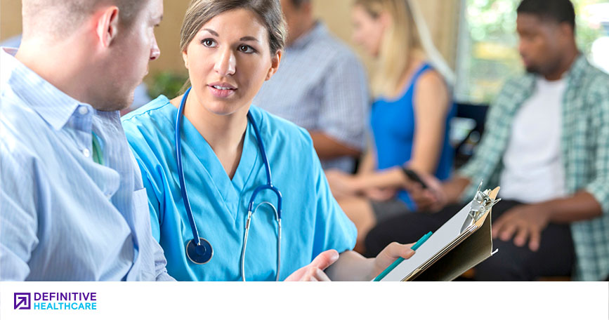 A female healthcare professional speaks with a patient in a doctor's office waiting room. Other patients wait patiently in the background.