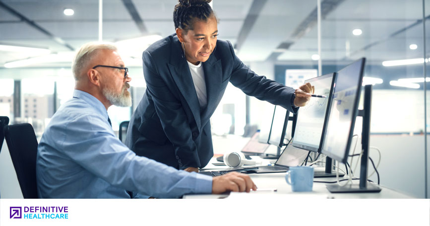 A woman in business attire talks with a seated man while gesturing toward a chart on his computer monitor.