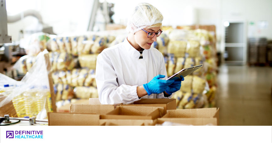 A person with glasses wearing a white jacket, hair net and blue gloves hold a tablet with boxes around them 