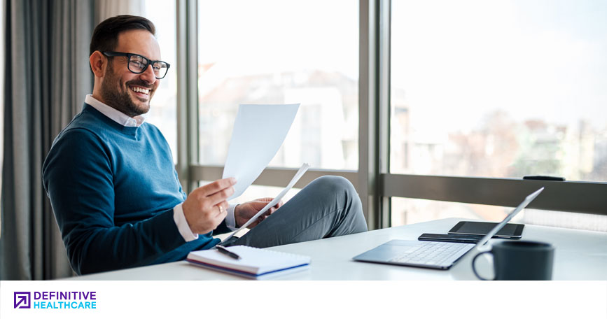 A business man with dark hair and glasses is sitting at a desk smiling at a piece of paper in his hand
