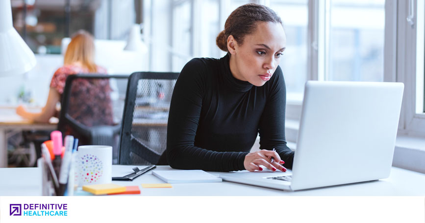 A women in a black turtle neck sitting at a desk looking at a white laptop