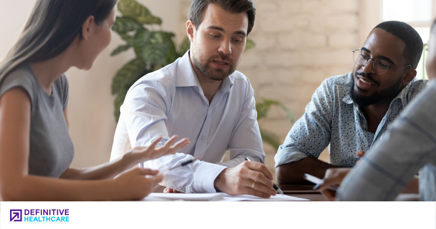 Three people wearing business attire speak together in a meeting room.