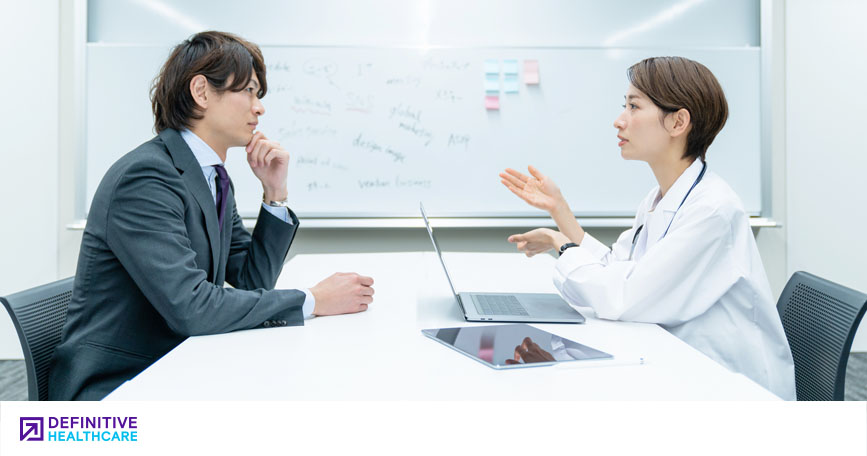 A clinician sits at a table with her laptop open and speaks to a medical affairs professional.