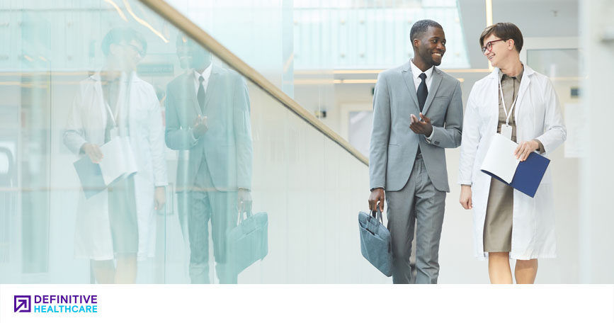 A clinician and a salesperson walk down a hospital hallway and talk while smiling.