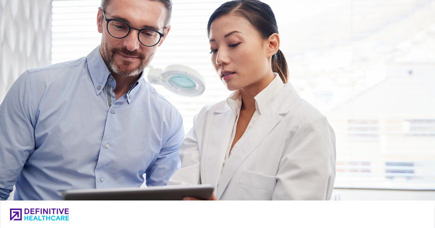 A man with glasses in a blue shirt stands next to a woman in a white medical jacket looking at a computer screen 