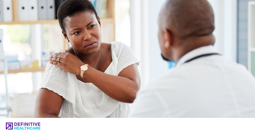 A female patient indicates to a male doctor that her shoulder is in pain.