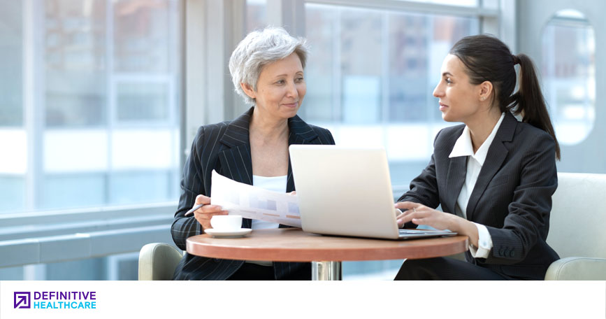 Two women in professional attire sit at a desk and review papers while one types on a laptop.