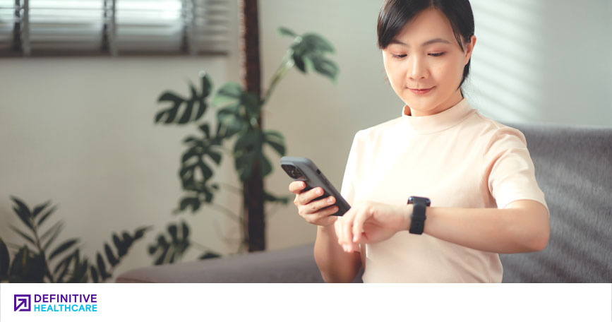 A woman looks at the health-monitoring watch on her wrist while holding a smartphone.