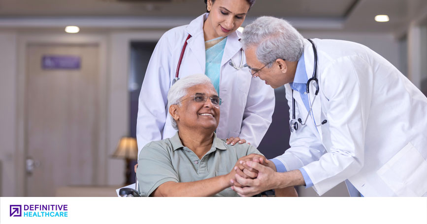 A doctor pushes a patient in a wheelchair while another doctor leans in to shake the patient's hand.