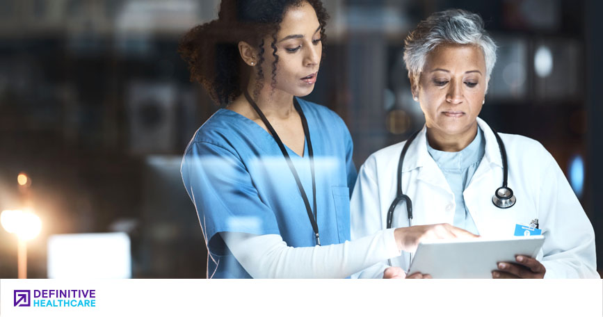 An image of a medium skinned woman with dark black hair in scrubs pointing at a chart held by a medium skinned woman with short silver hair in a white lab coat