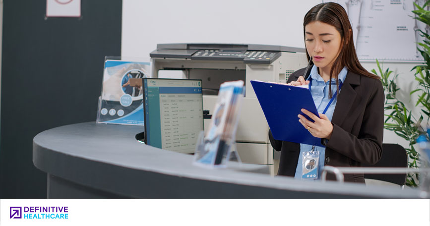 A healthcare professional sits behind a desk and reviews information on a clipboard.