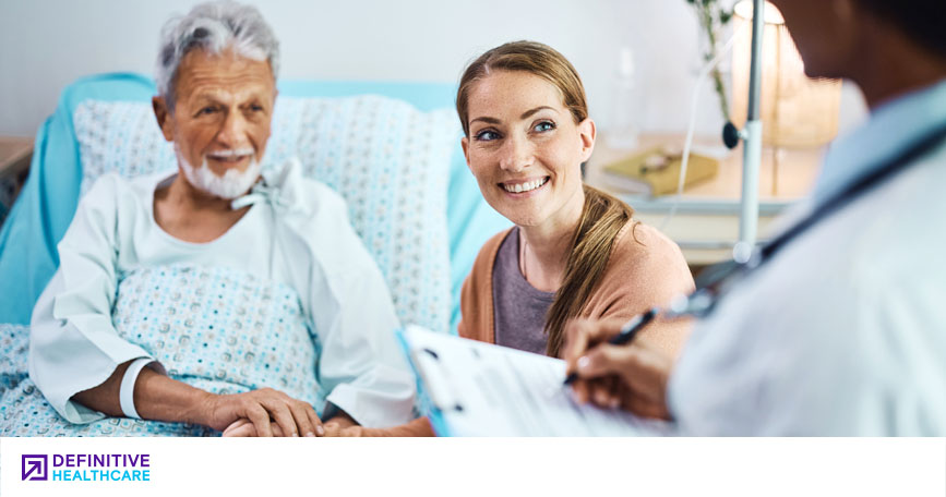 A patient sits upright in bed with his loved one nearby as a doctor speaks with them.