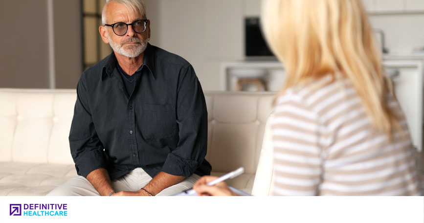 A man sits on a couch while a behavioral health professional takes notes on her notepad.