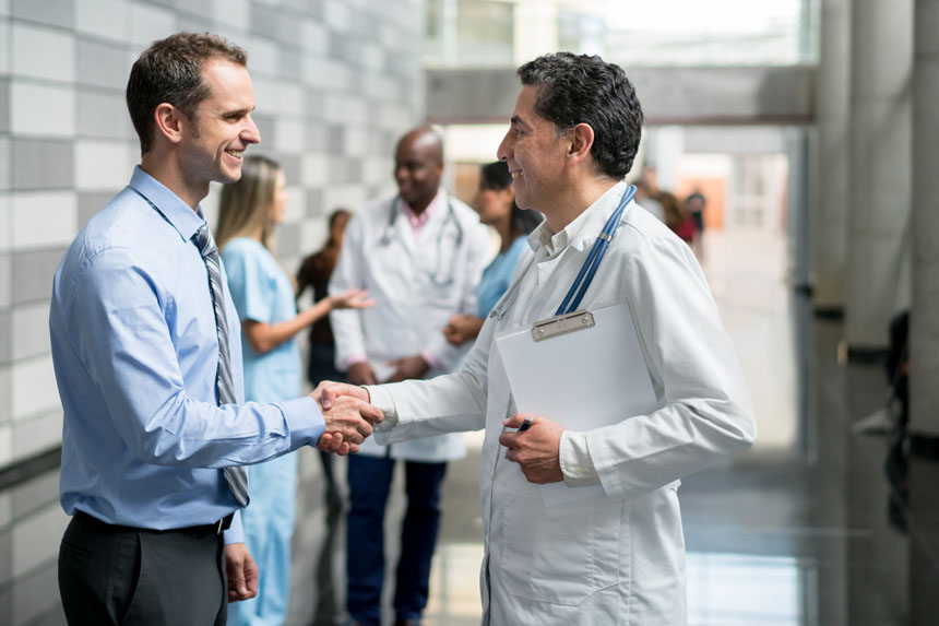 A man in business attire shakes hands with a male doctor in a hospital hallway. Other healthcare professionals talk in the background. 