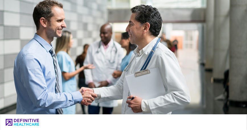 A man in business attire shakes hands with a male doctor in a hospital hallway. Other healthcare professionals talk in the background.