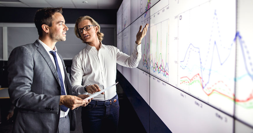 Two men in business attire look at a chart projected onto a wall. They talk as one of the men gestures to the data.