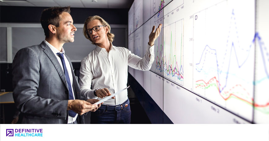 Two men in business attire look at a chart projected onto a wall. They talk as one of the men gestures to the data.