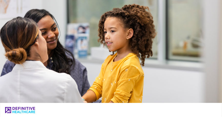 A young girl sits on an examination table in a medical office accompanied by her mother and a female doctor.