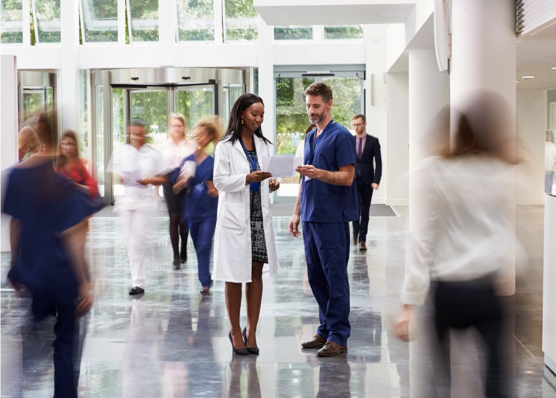 A woman healthcare professional is looking at papers with a male healthcare professional in a lobby. Other people are walking around them in the background.