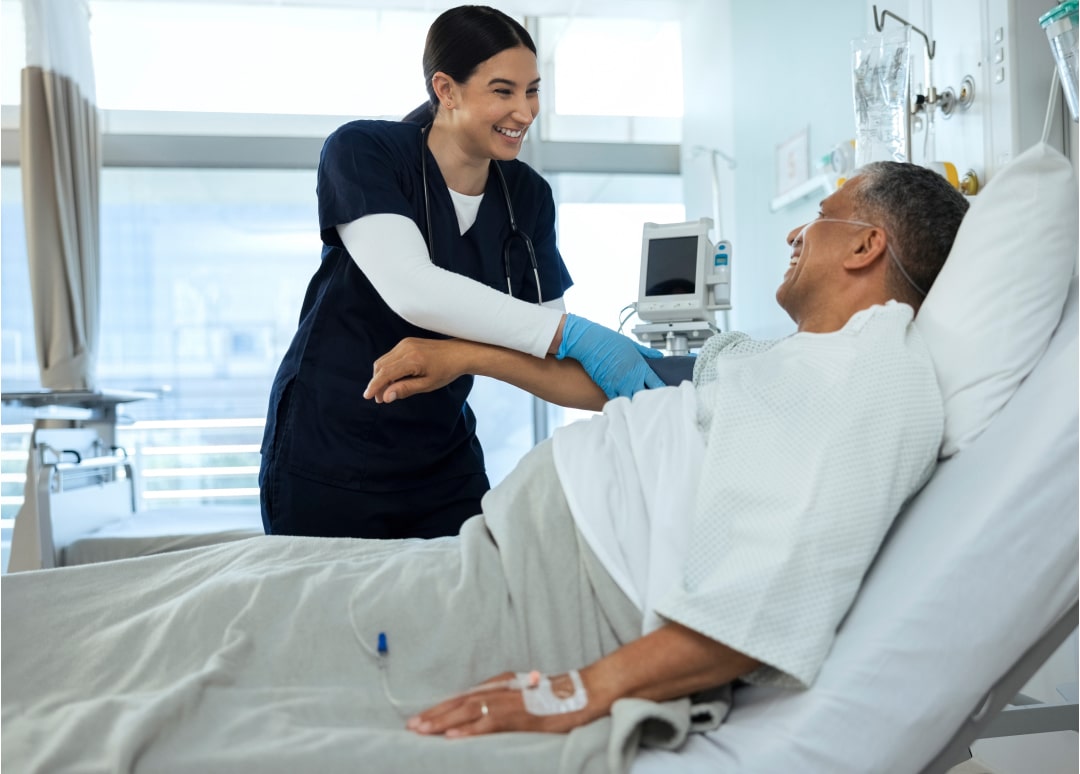 A healthcare professional smiles while holding the arm of a patient who is smiling as he sits in a hospital bed.