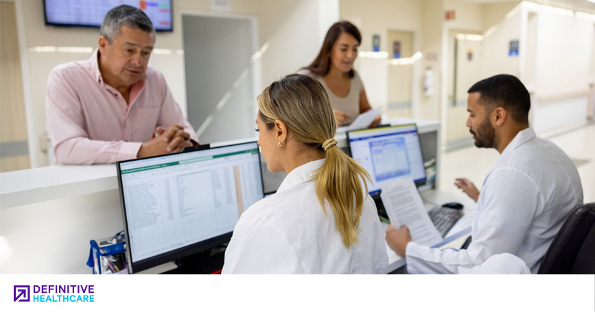 Two medical administrative personnel sit at computer workstations behind a counter in a healthcare facility, helping two patients with billing.