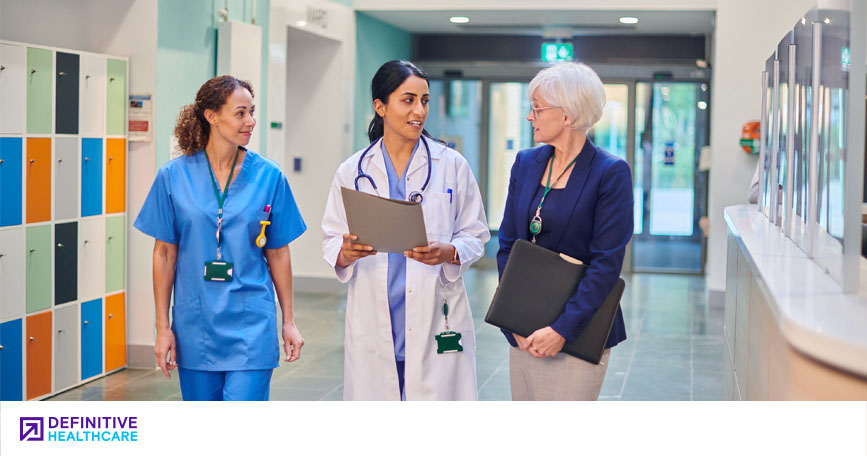 Three female healthcare professionals smile and talk as they walk through a well-lit, modern hospital hallway.