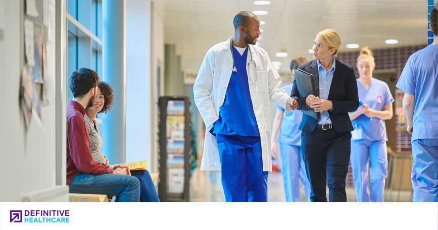 A busy hospital hallway. Two visitors sit and chat on a bench in front of a window, while a doctor talks with a medtech representative. Nurses can be seen walking up and down the hall.