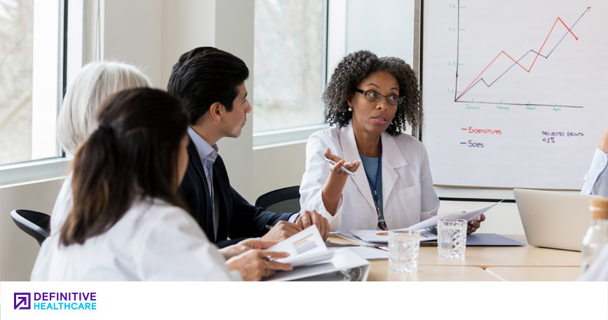 Several healthcare professionals and marketing professionals sit around a conference table and talk while reviewing data. A whiteboard in the background shows a line chart.