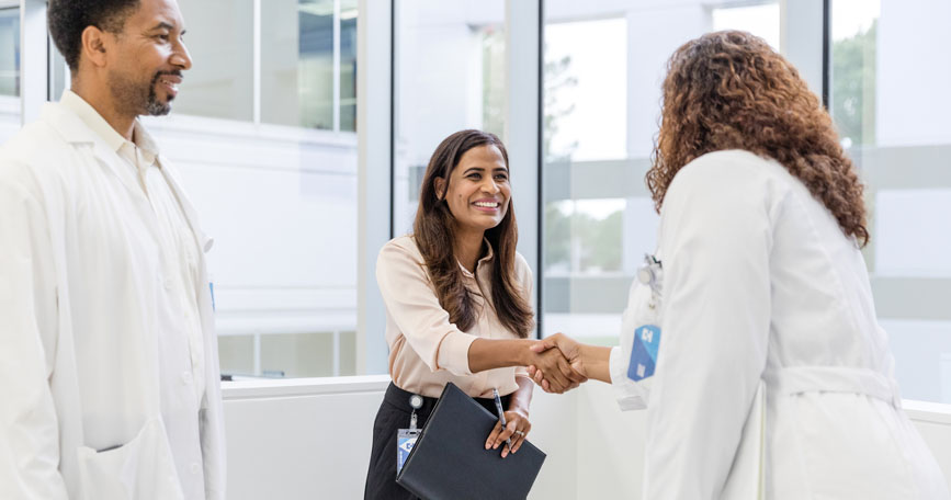 A sales professional shakes hands with a physician while another medical professional looks on.