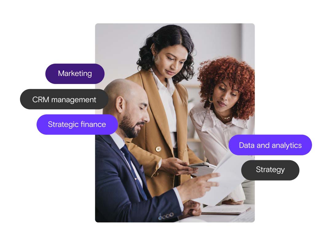 A man in business attire sits holding a paper while two women, also in business attire, one standing, the other leaning on the table look down at the paper.