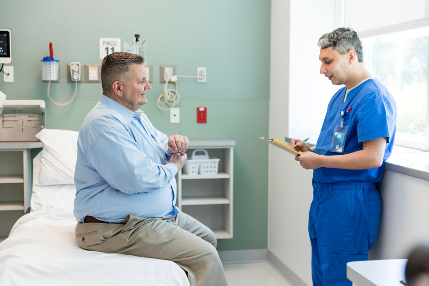 A heavyset male patient sits on an exam room table and talks with a healthcare professional who's reviewing a chart.