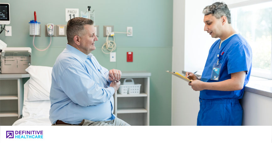 A heavyset male patient sits on an exam room table and talks with a healthcare professional who's reviewing a chart.
