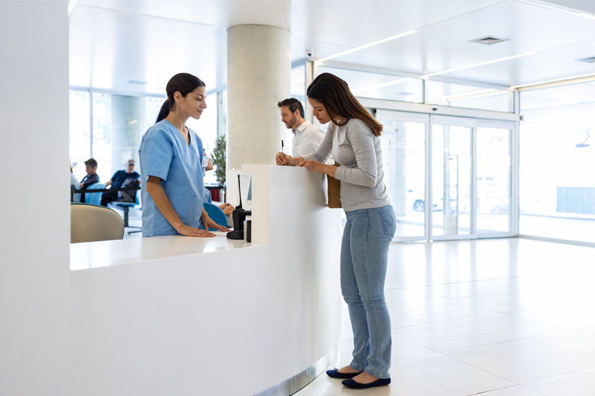 A female patient fills out a form at the front desk of a medical facility while a healthcare professional looks on.