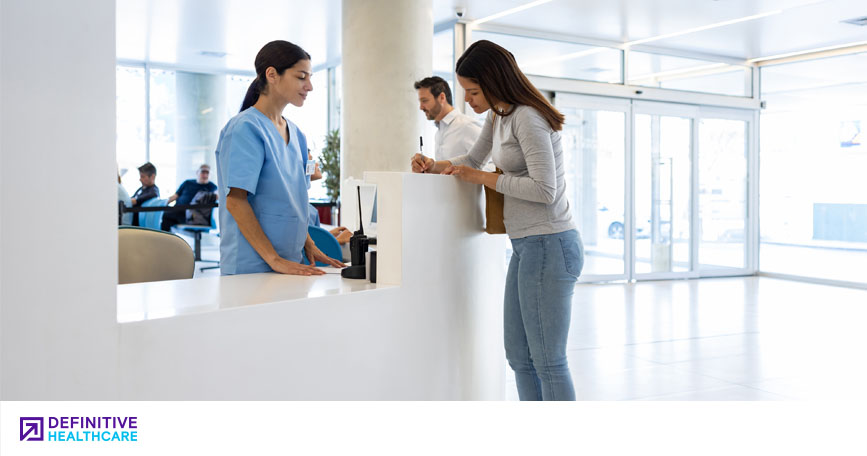 A female patient fills out a form at the front desk of a medical facility while a healthcare professional looks on.
