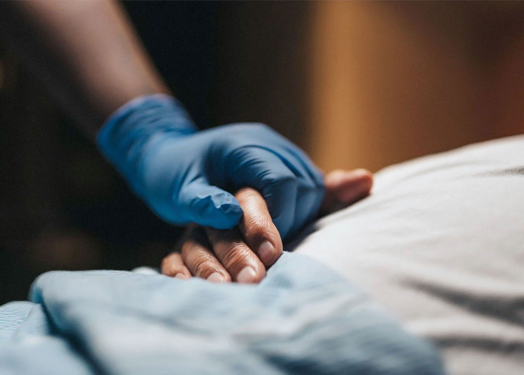 aA woman and a man dressed in business attire are having a conversation. The woman is walking while looking at the man who is in a wheelchair and has a laptop in his lap. A healthcare professional is standing in the background.