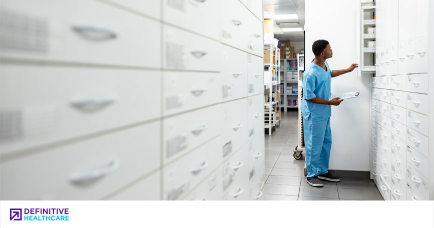 A healthcare worker stands amid rows of shelves and drawers in a supply room and examines the contents of a cabinet.
