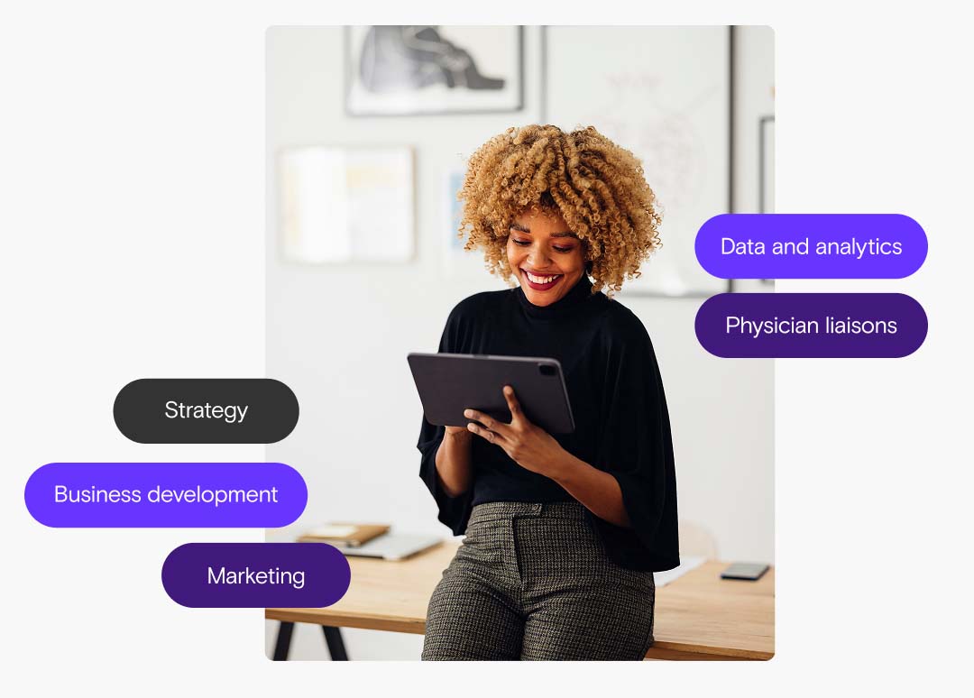 A woman in business casual attire leans against a table smiling while looking down at a tablet she is holding.