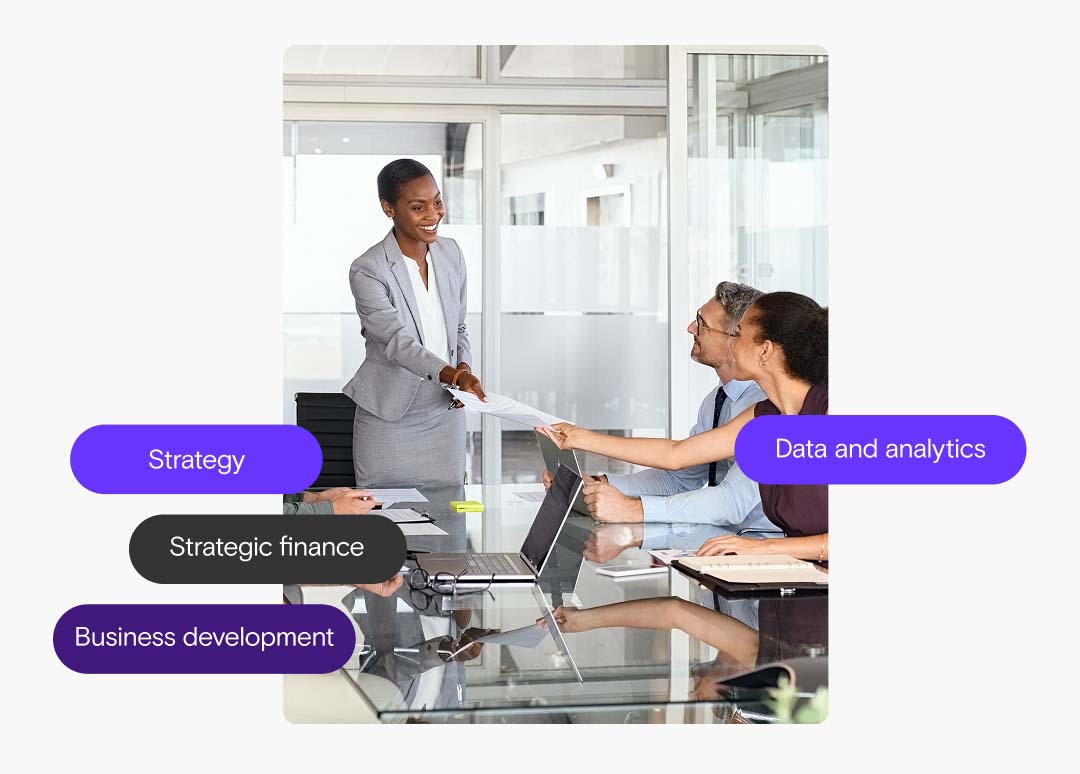 A woman in business attire stands and smiles while holding a paper during a meeting. Another woman holds the other end of the paper while sitting.