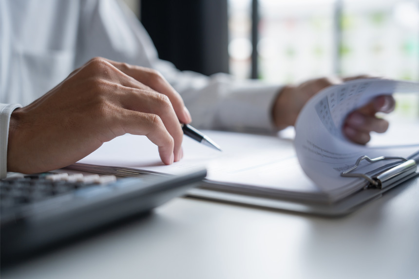 A close-up shot of a person's hands. The left hand flips through forms on a clipboard while the right hand holds a pen.