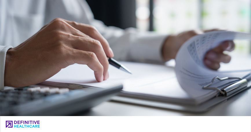 A close-up shot of a person's hands. The left hand flips through forms on a clipboard while the right hand holds a pen.