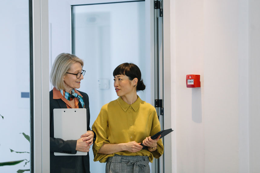 Two females dressed in business casual attire, one holding a clipboard, the other a tablet, talk while standing in a doorway.