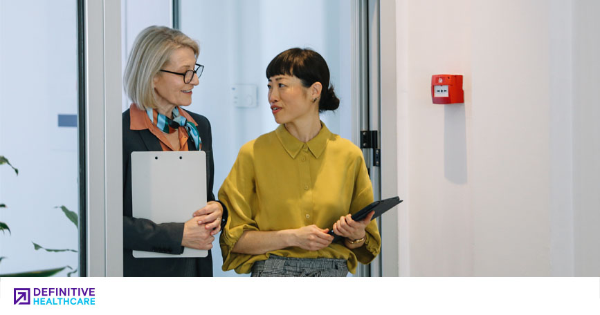 Two females dressed in business casual attire, one holding a clipboard, the other a tablet, talk while standing in a doorway.