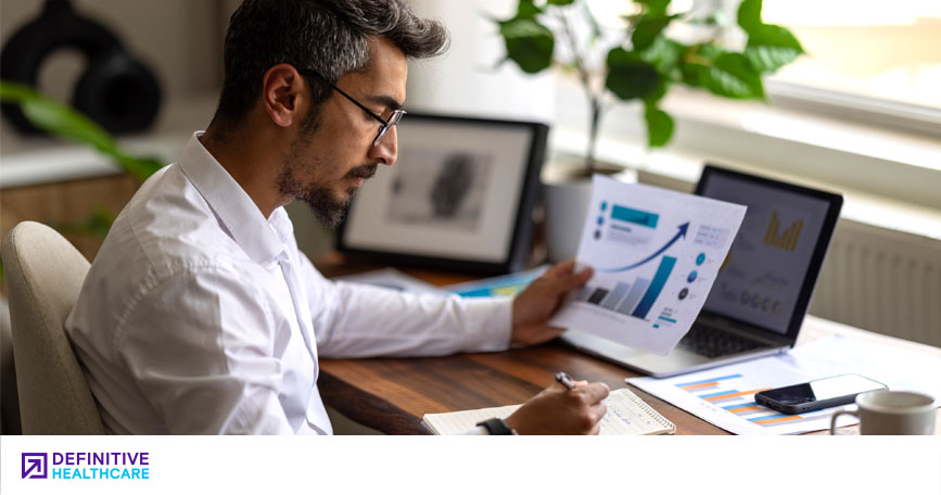 A man in business attire sits at a desk and writes on a notepad while holding up a piece of paper.
