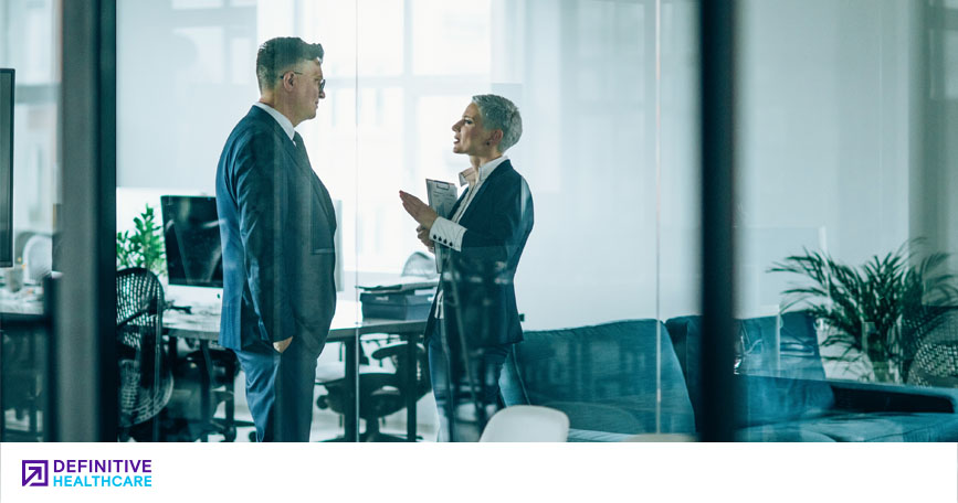 Two people dressed in business attire are standing up having a conversation in an office setting.