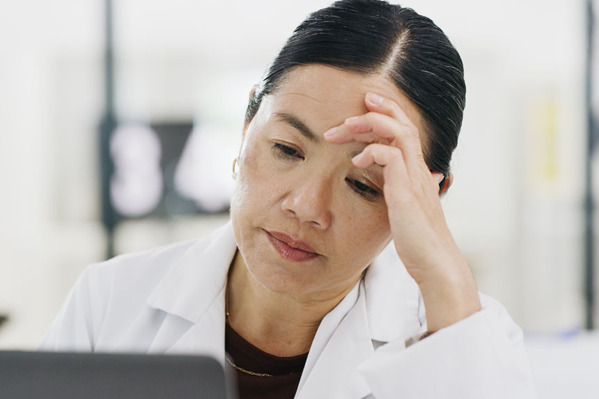 A female healthcare professional in a white coat presses a hand to her temple and appears worried.