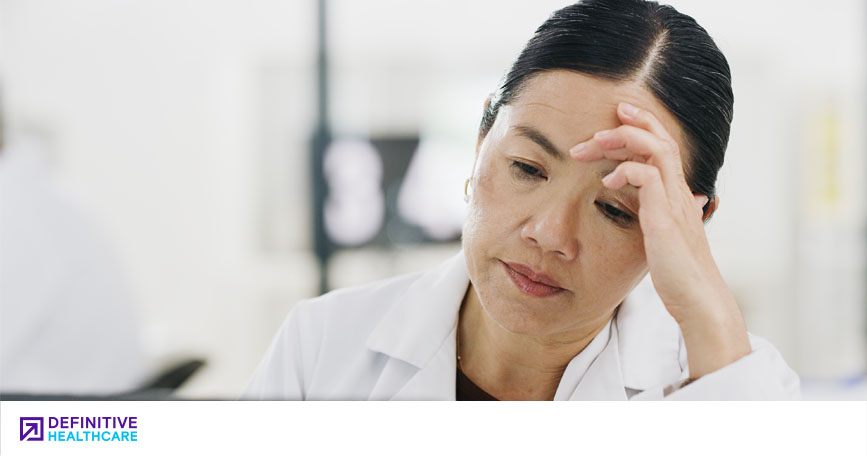 A female healthcare professional in a white coat presses a hand to her temple and appears worried.