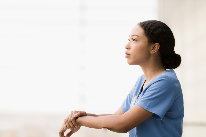 A female healthcare professional leans against a railing and looks thoughtful.