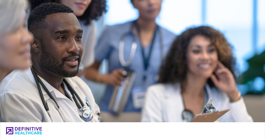 Several smiling physicians sit and stand around a table and listen to a speaker (out of frame).